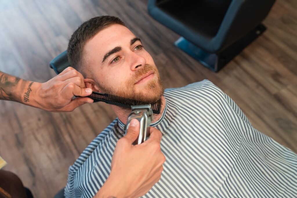 young barber shaving male customers beard with comb hair clipper barbershop explaining if do barbers cut long hair