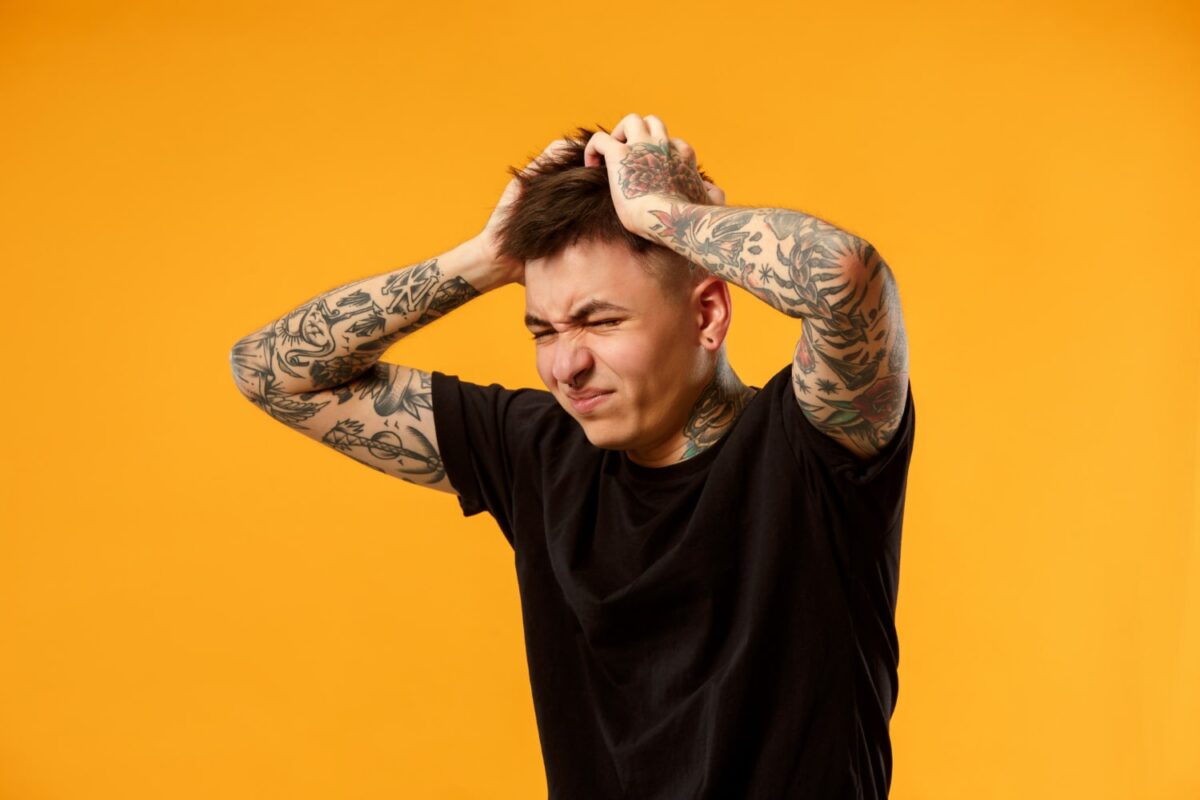 young man with tattoos on his arms touching his head with both hands angry after receiving a bad haircut